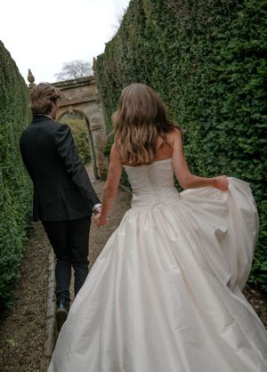 A couple walks away from the camera through a garden path lined with lush green ivy walls and stone architecture.