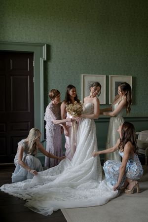 A sequence of wedding preparation photos showing bridesmaids helping adjust a flowing lace wedding dress train in a room.