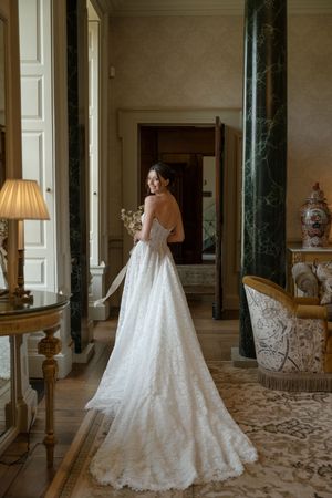 Elegant white lace wedding gown with cathedral train displayed in a luxurious room with ornate decor and columns.
