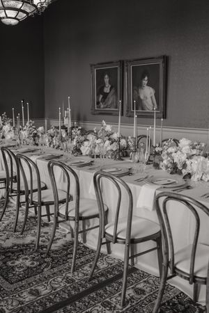 Black and white photo of an elegant wedding reception setup with long tables and chairs in a formal dining room.