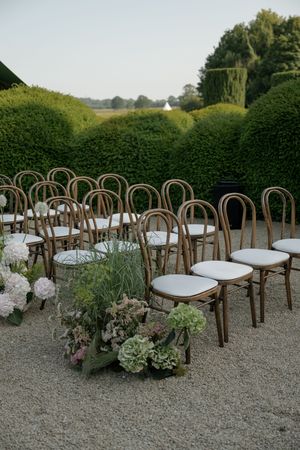 Wooden bentwood chairs with white cushions arranged outdoors on gravel with elegant floral arrangements for a garden event.