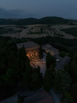 Aerial twilight view of a Tuscan villa surrounded by cypress trees and rolling hills, lit by warm string lights.