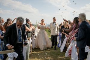 Bride and groom walk down aisle as guests toss confetti at outdoor wedding ceremony with scenic view.