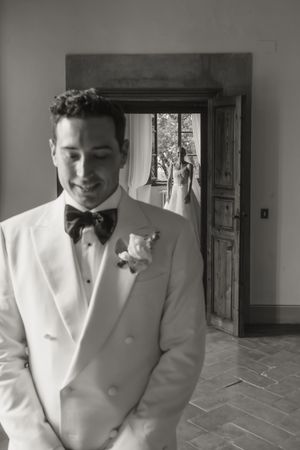 Groom in white tuxedo with boutonniere smiles, bride in wedding dress visible through doorway behind him.