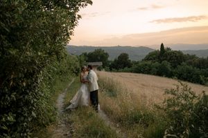 Bride and groom embracing on a rustic path at sunset with rolling Tuscan hills and golden fields in the background.