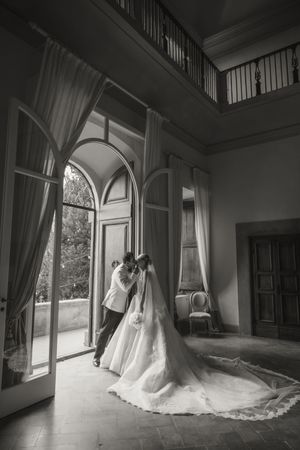 Bride and groom share romantic embrace in grand villa ballroom with arched windows in black and white.