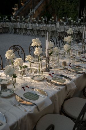 Elegant outdoor wedding reception table with white roses, taper candles, crystal glassware, and fine china on white linen.
