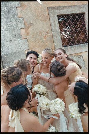 Bride surrounded by bridesmaids in champagne dresses sharing a joyful group hug with white bouquets.
