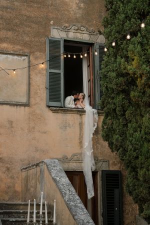 Bride and groom sharing a romantic kiss in a rustic Italian villa window, adorned with string lights and a flowing veil.