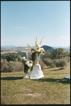 Elegant white floral arrangements on draped pedestals set against rolling Tuscan hills under a clear blue sky.