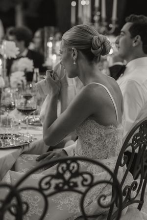 Black and white photo of an elegant bride in lace dress seated at a wedding reception dinner table with wine glasses.