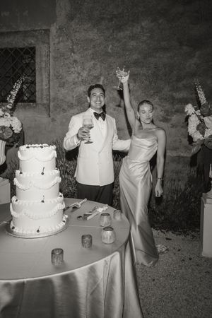 Bride and groom toast with champagne beside a white tiered wedding cake in a black and white photo.