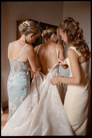 Two bridesmaids help a bride fasten her lace wedding gown from behind in a warmly lit bridal suite.