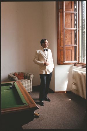 A groom in a white tuxedo jacket and bow tie stands by a window near a billiard table in an elegant room.