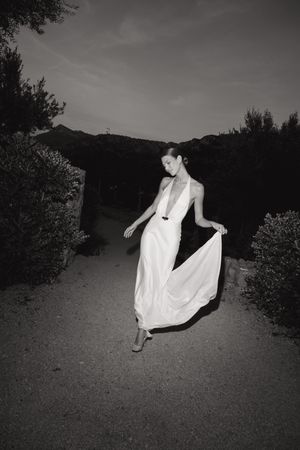 A graceful figure in a flowing white dress dances on a path at dusk in a black and white photograph.