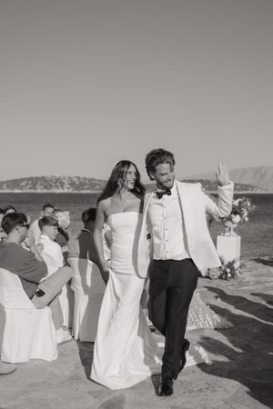 Wedding guests in formal attire gather on a sunny beach with white chair covers arranged for an outdoor ceremony.