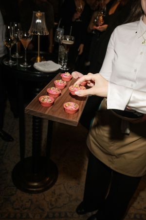 Server carrying wooden tray with pink desserts at an evening event.