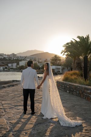 A couple in wedding attire walks along a stone path at sunset with palm trees and mountains in the background.