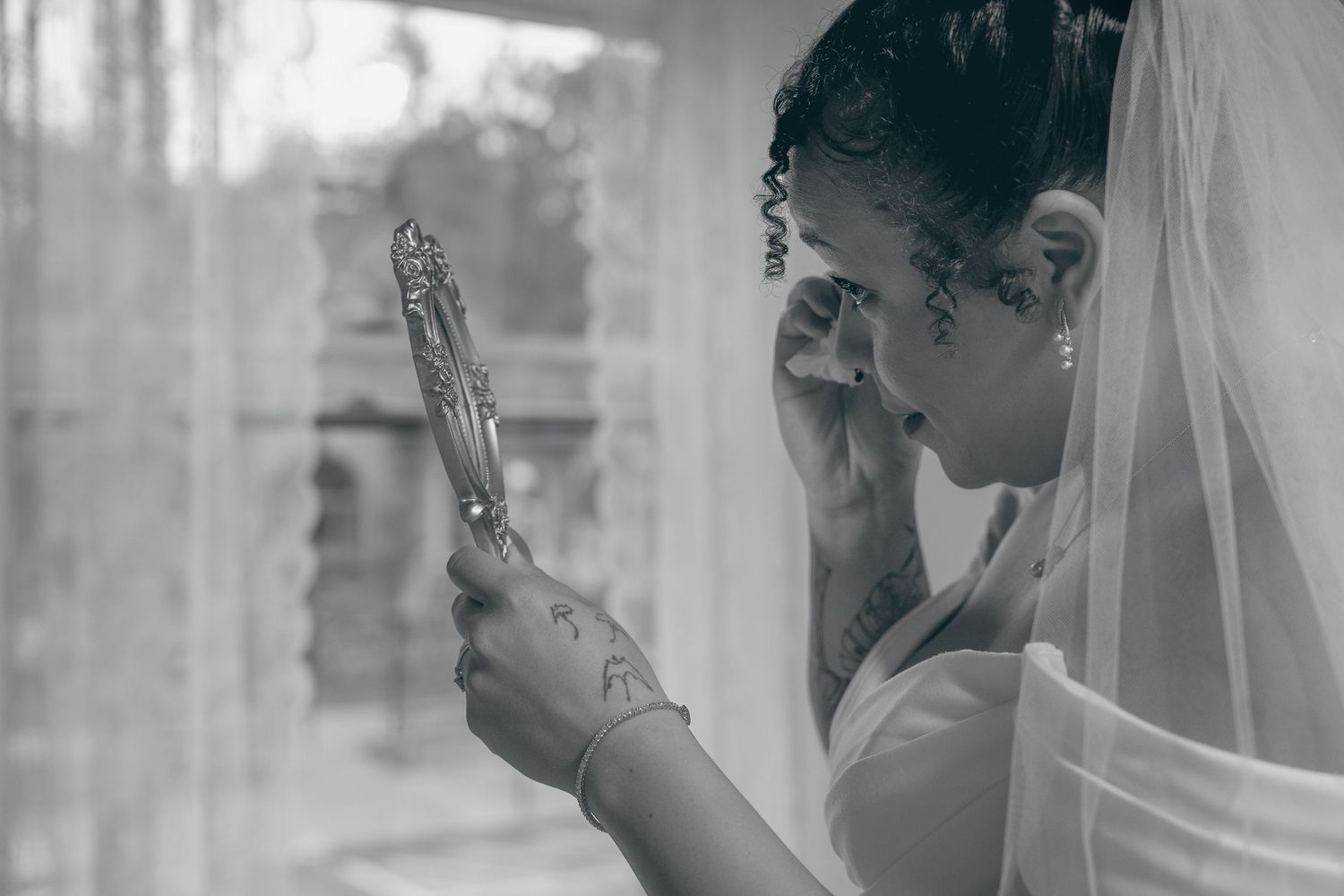 A bride in a white veil looks at her reflection in an ornate handheld mirror in a black and white photograph.
