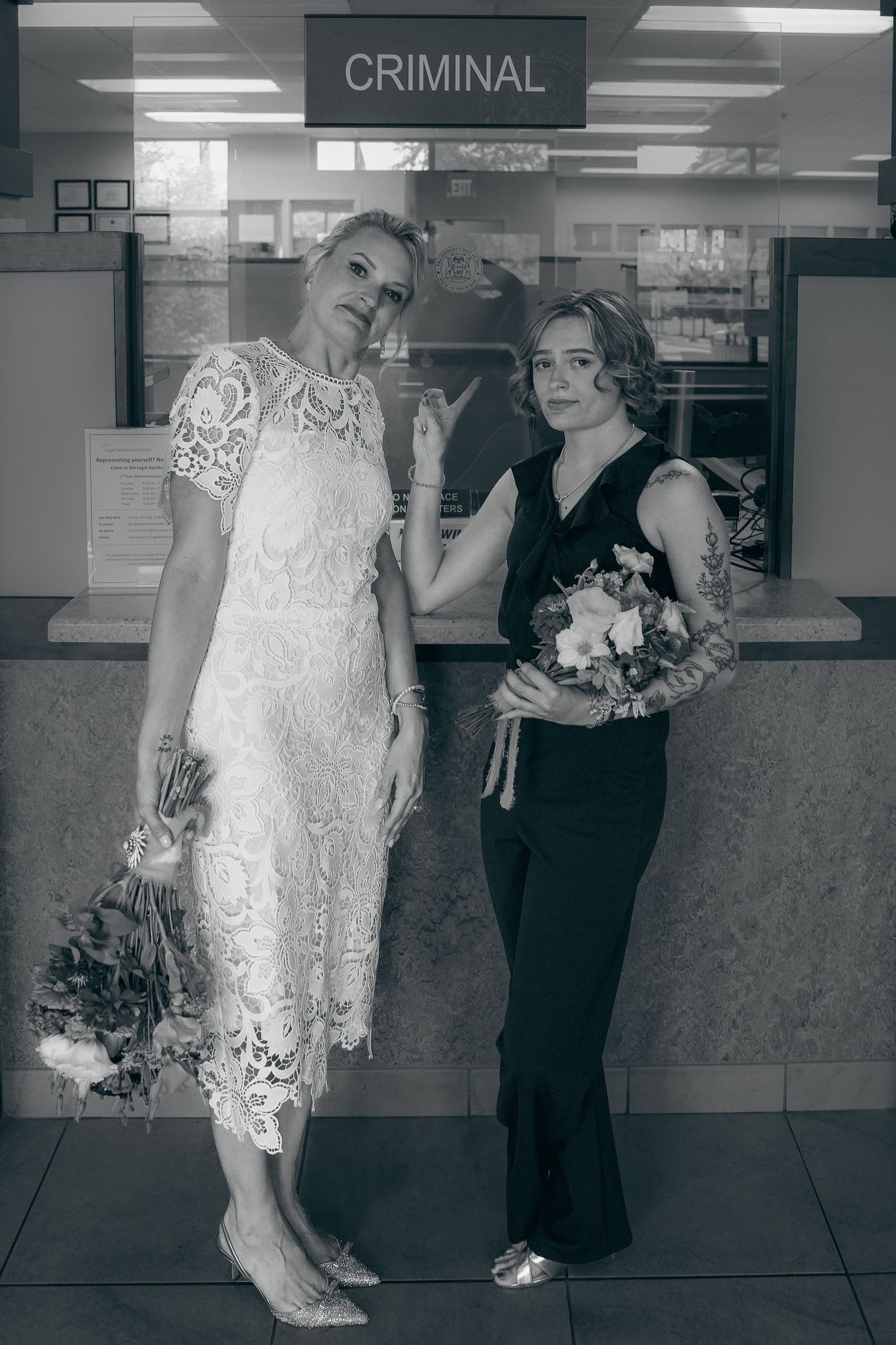 Two people pose in formal attire in front of a window marked 'CRIMINAL' in a black and white photo.