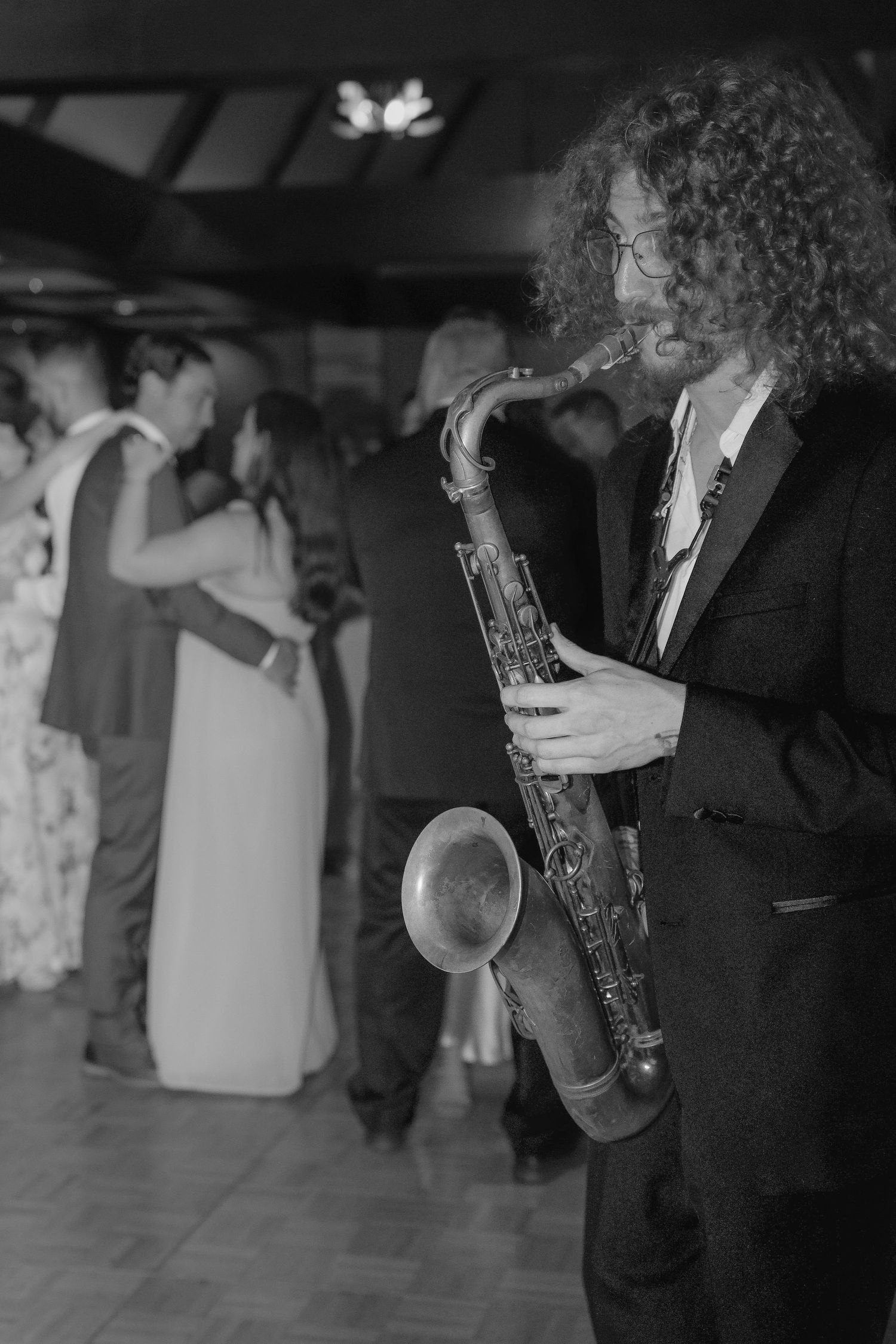 A saxophone player in a suit performs at an event while guests dance in the background.