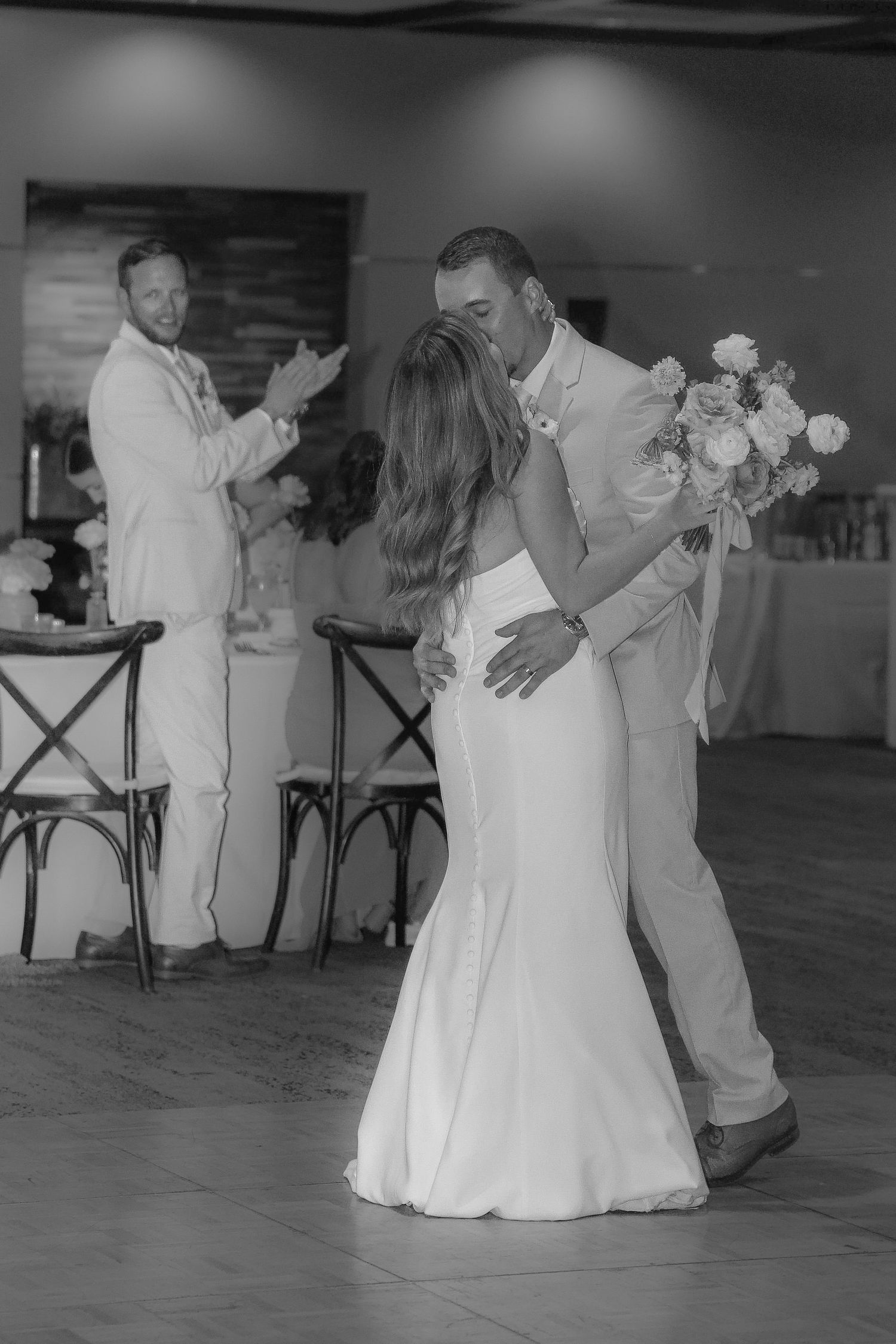 A couple shares their first dance at a wedding reception while a guest in white looks on from the background.