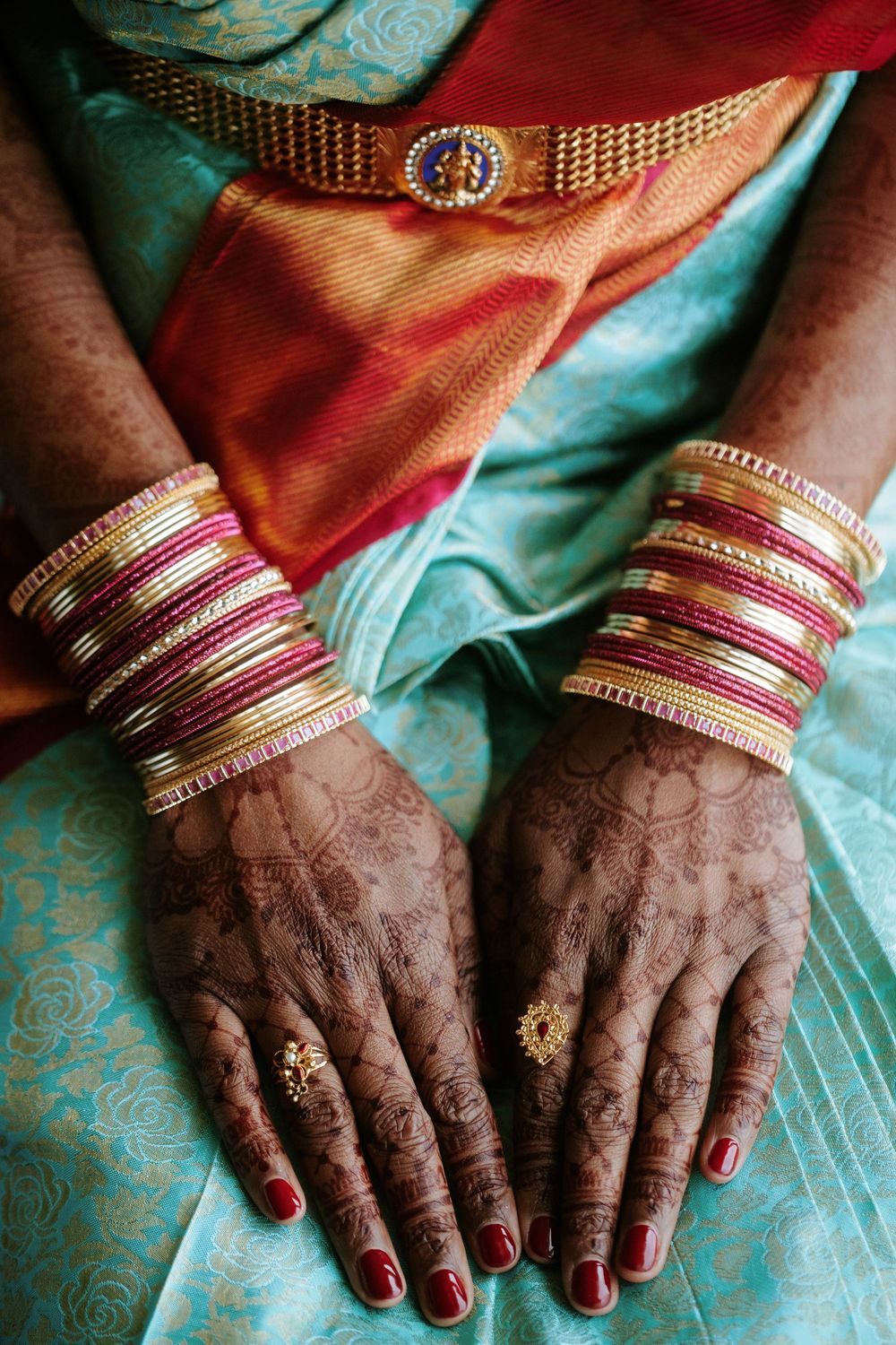 Close-up view of hands adorned with intricate henna designs and pink bangles against turquoise fabric.