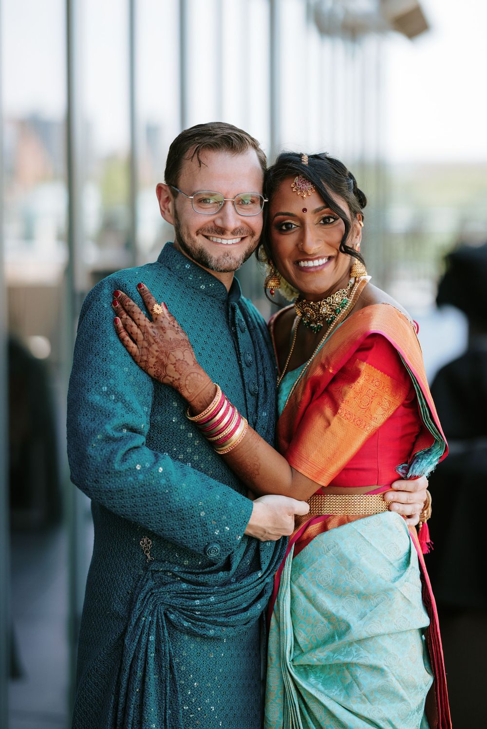 A couple embraces at a wedding celebration, with one wearing a turquoise outfit and the other in traditional South Asian attire.