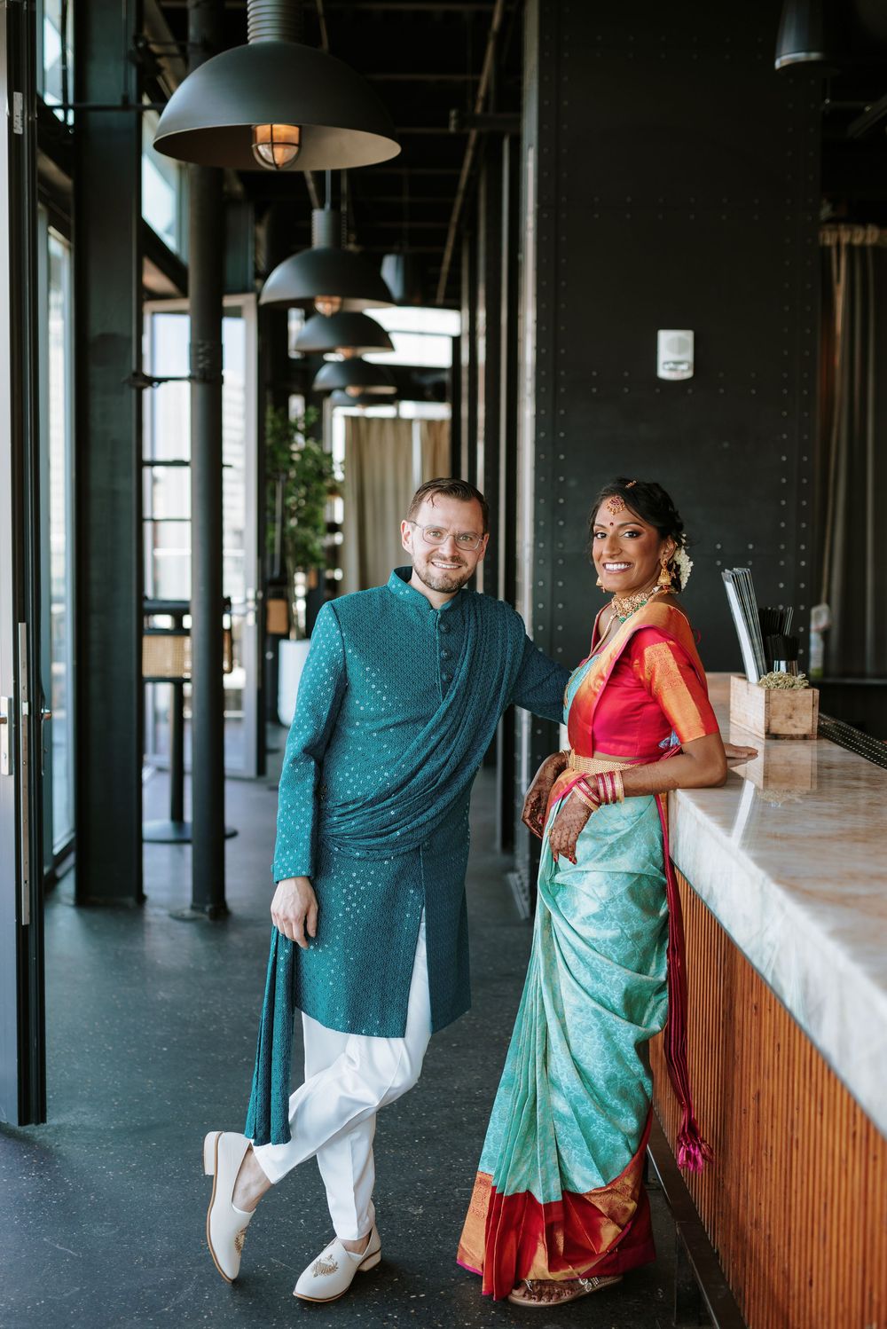 Couple in traditional Indian attire dancing and posing playfully in a modern industrial-style hallway.