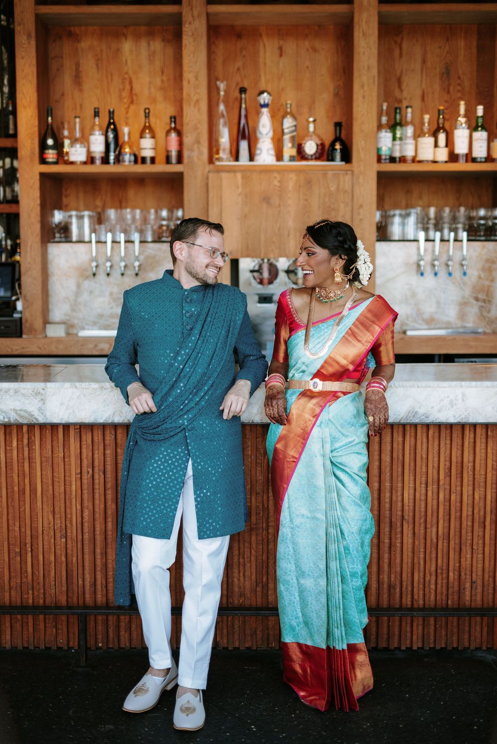 A couple poses playfully in front of a wooden bar with shelves of bottles in the background.