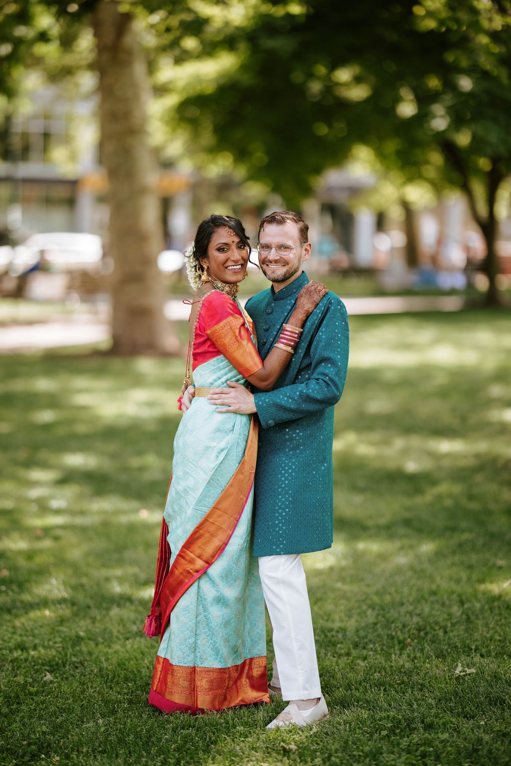 A couple in traditional Indian and Western attire embrace joyfully in a sunny park setting.