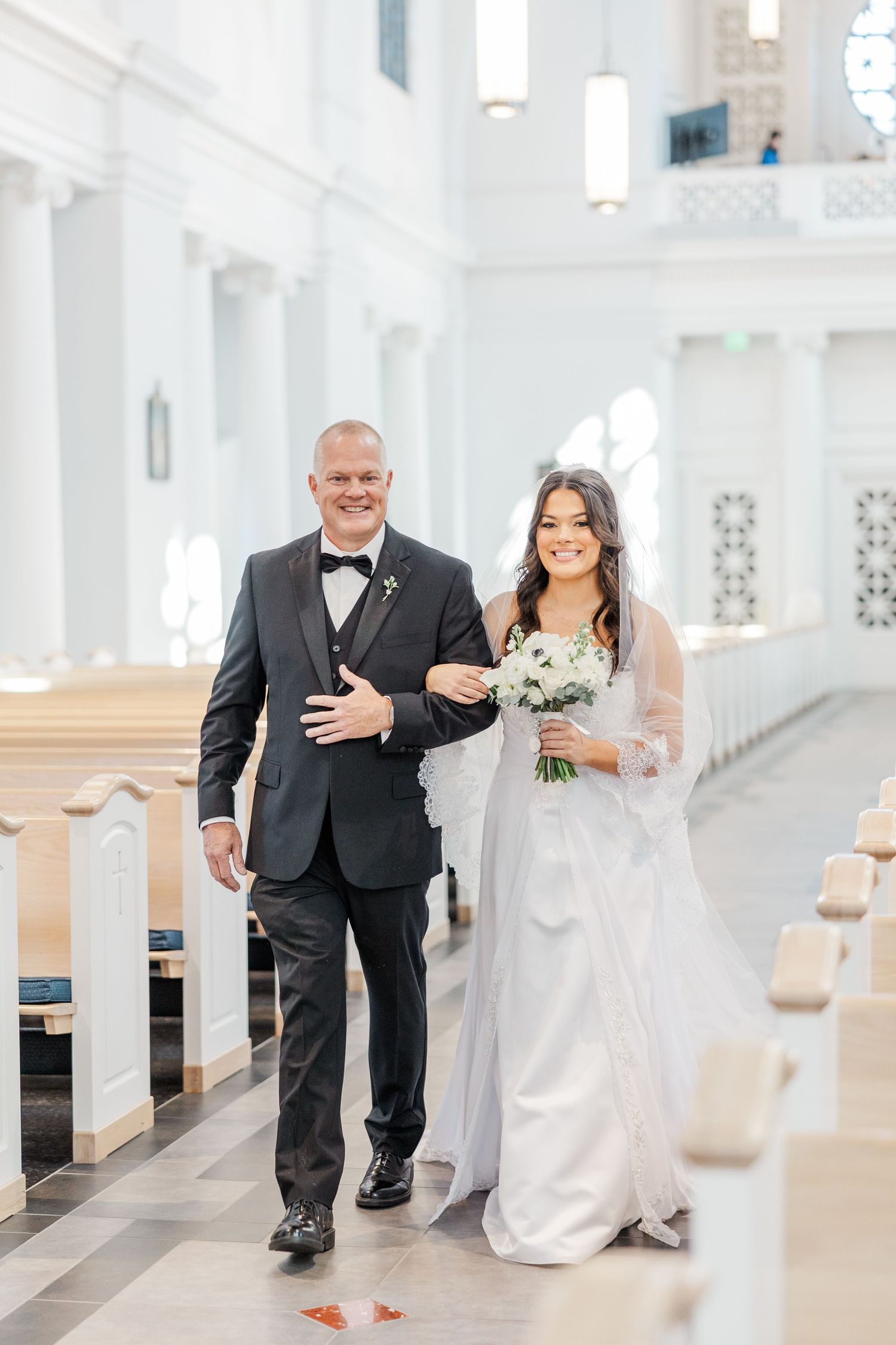 A bride in a white wedding dress and veil walks down a church aisle with an escort in a black tuxedo.