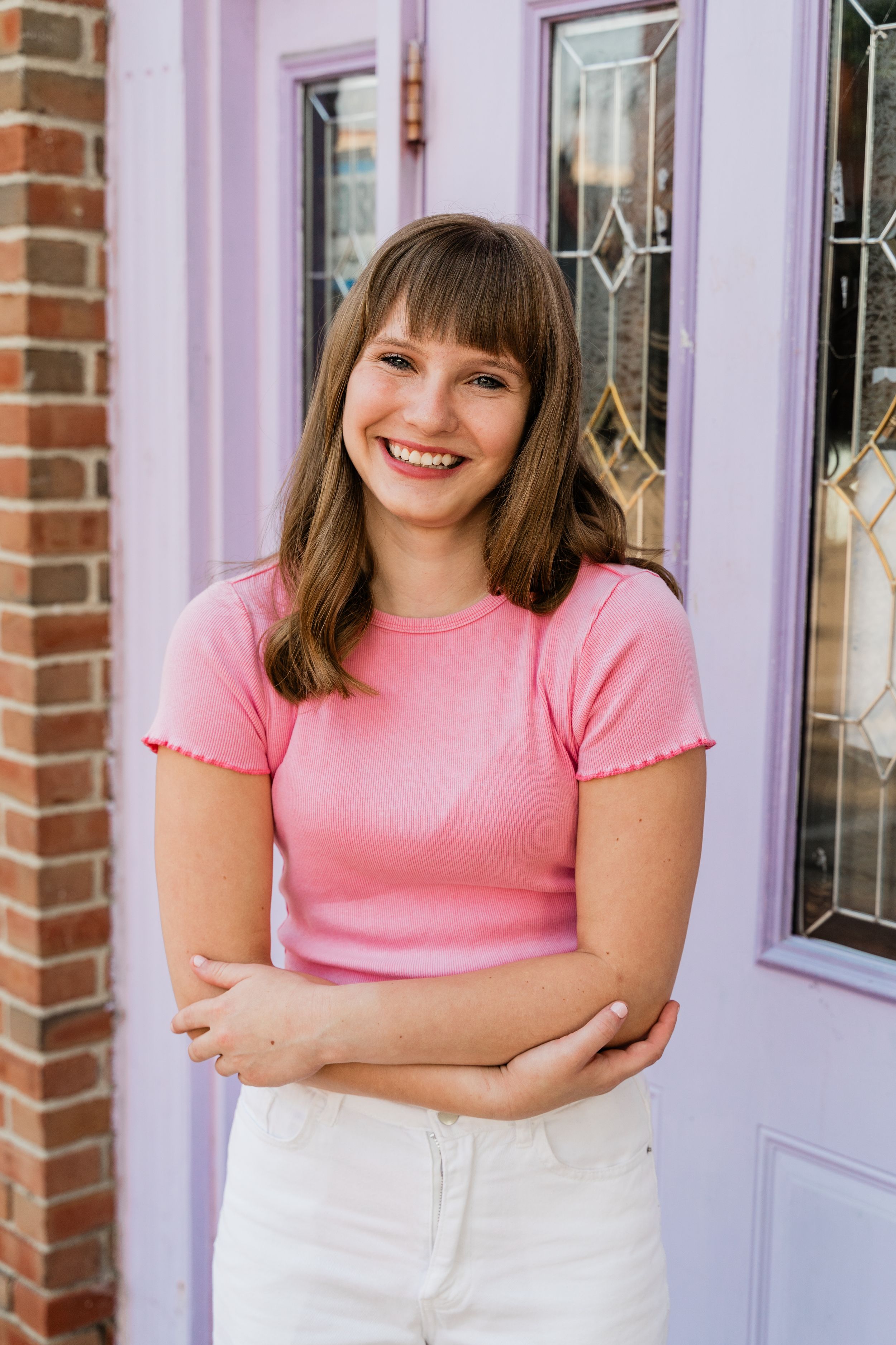A smiling person wearing a pink t-shirt and white pants stands against a purple wall with brick accents.