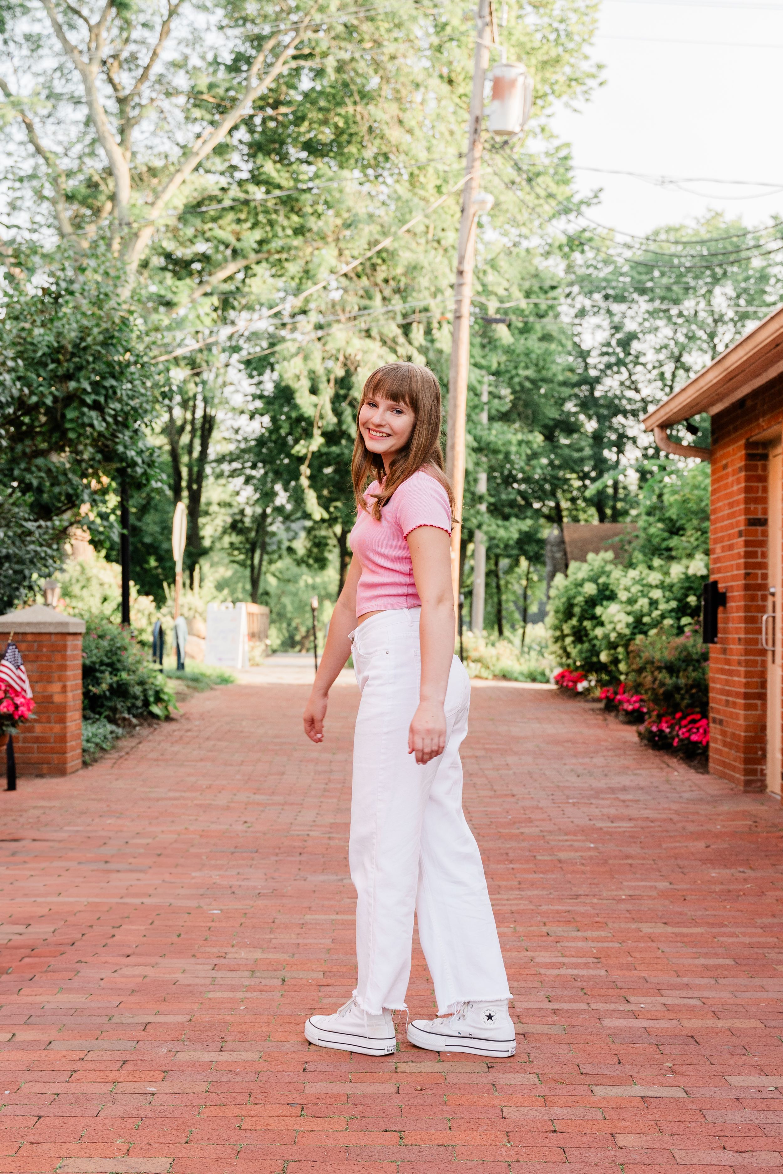 A young girl in a pink shirt and white pants poses on a brick walkway near a wooden structure.