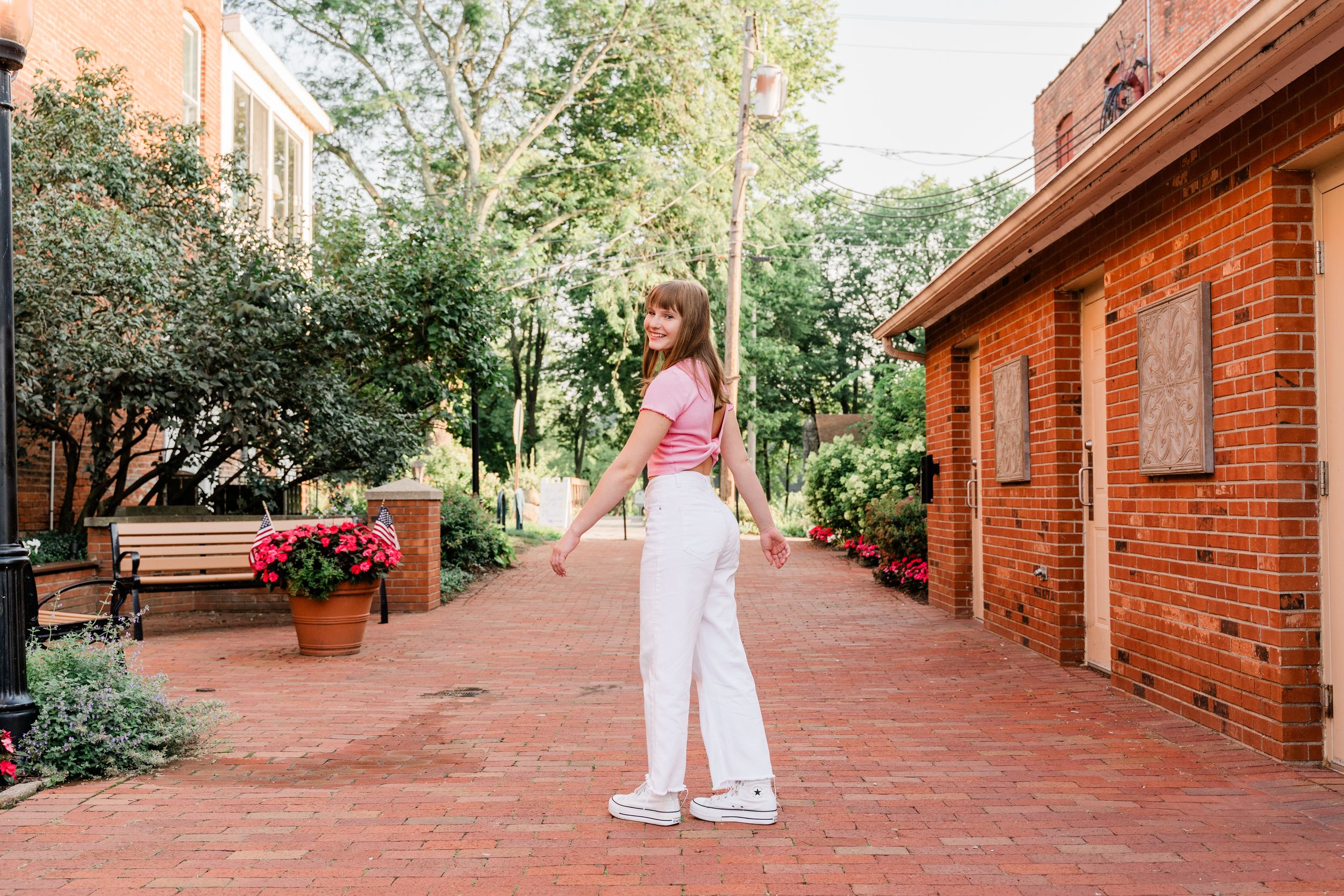 Someone in white pants and pink top stands on red brick walkway surrounded by lush greenery and brick buildings.