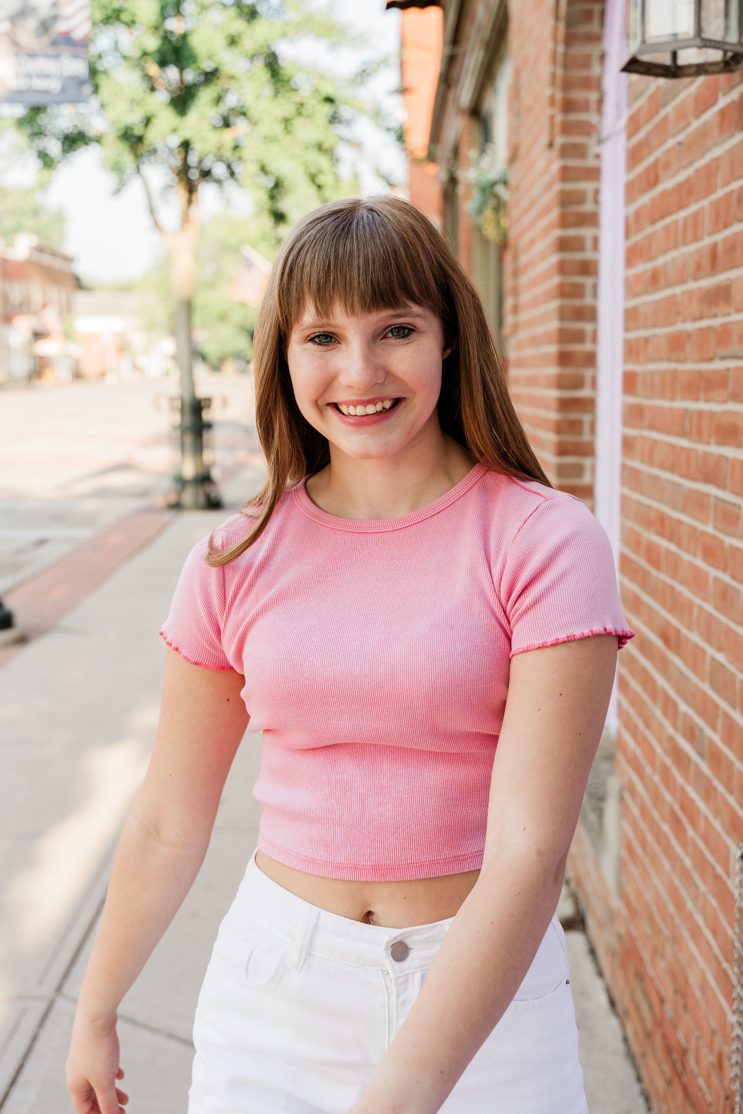 Person wearing a pink crop top and white pants poses against a brick wall in an outdoor setting.