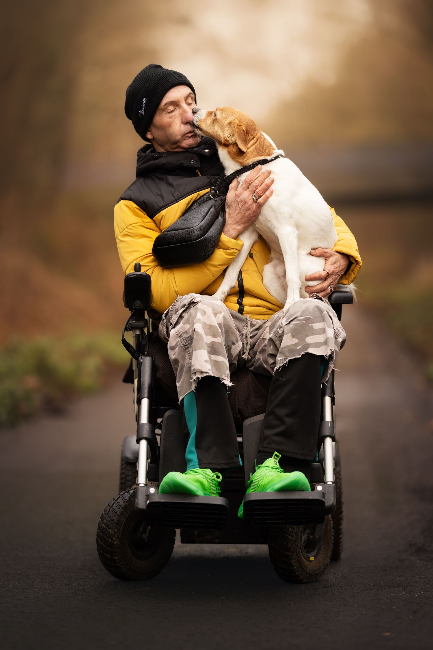 A person in a wheelchair wearing a yellow jacket and black beanie shares a tender moment with a small dog on their lap.