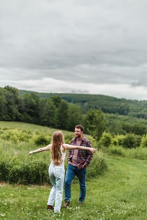 A couple spins and dances together in a flowering meadow surrounded by green hills and forests.