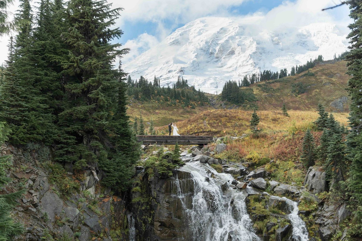 Scenic mountain waterfall cascades down rocky cliffs surrounded by evergreen trees and autumn foliage in the wilderness.