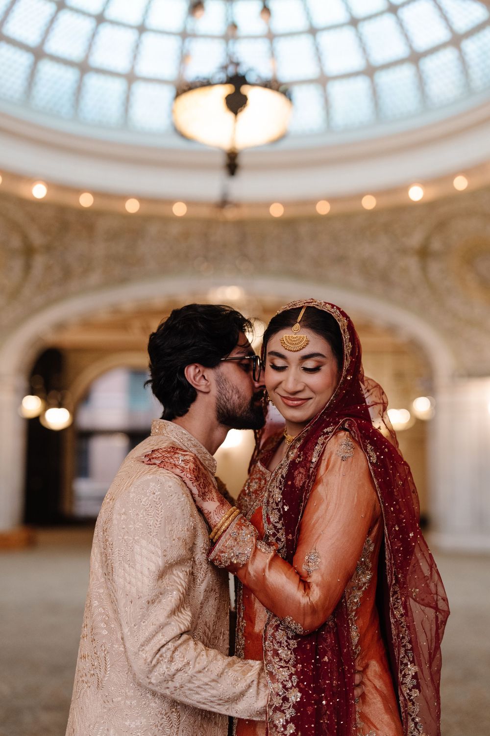South Asian bride and groom in traditional wedding attire embracing under ornate domed ceiling.