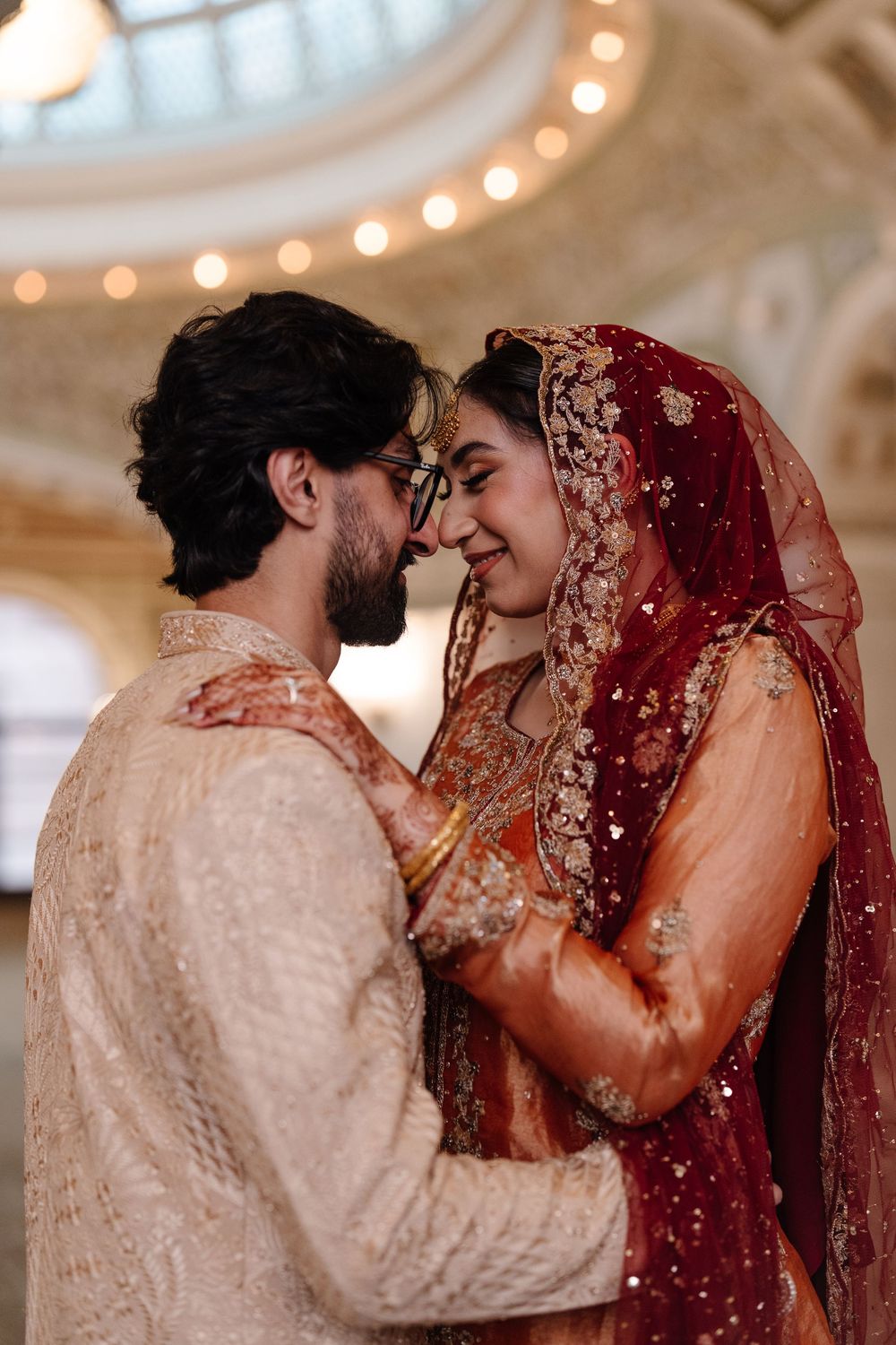 South Asian bride and groom in traditional wedding attire sharing romantic moment under glowing domed ceiling.