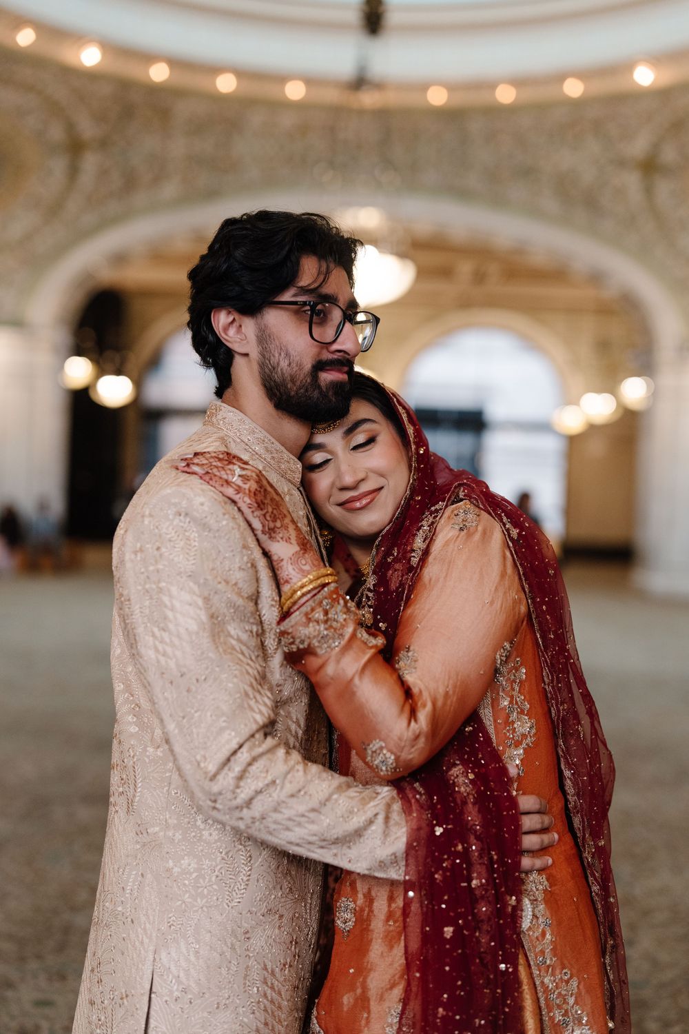 South Asian wedding couple embracing in grand hall, bride in red dupatta smiling while groom holds her close.