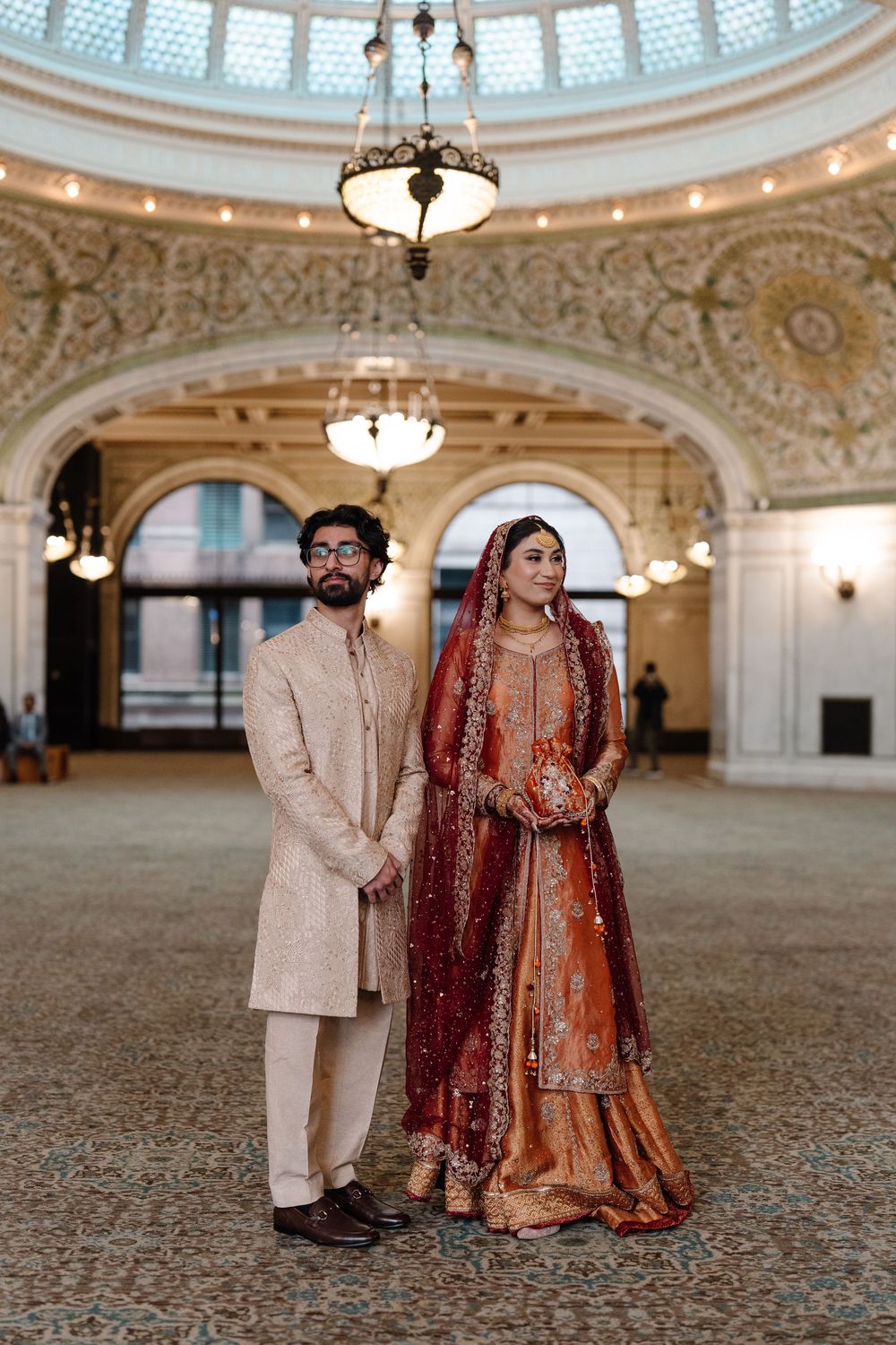 South Asian couple in traditional wedding attire standing under ornate domed ceiling of elegant wedding venue.