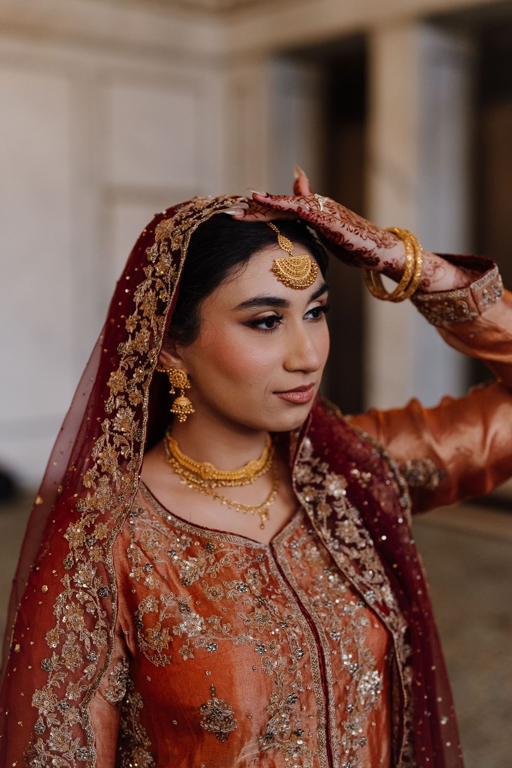 South Asian bride in rust orange bridal lehenga with gold jewelry and red dupatta posing elegantly.