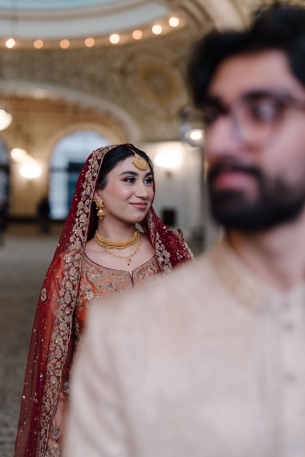 South Asian bride in red and gold bridal attire smiling softly with groom in foreground.