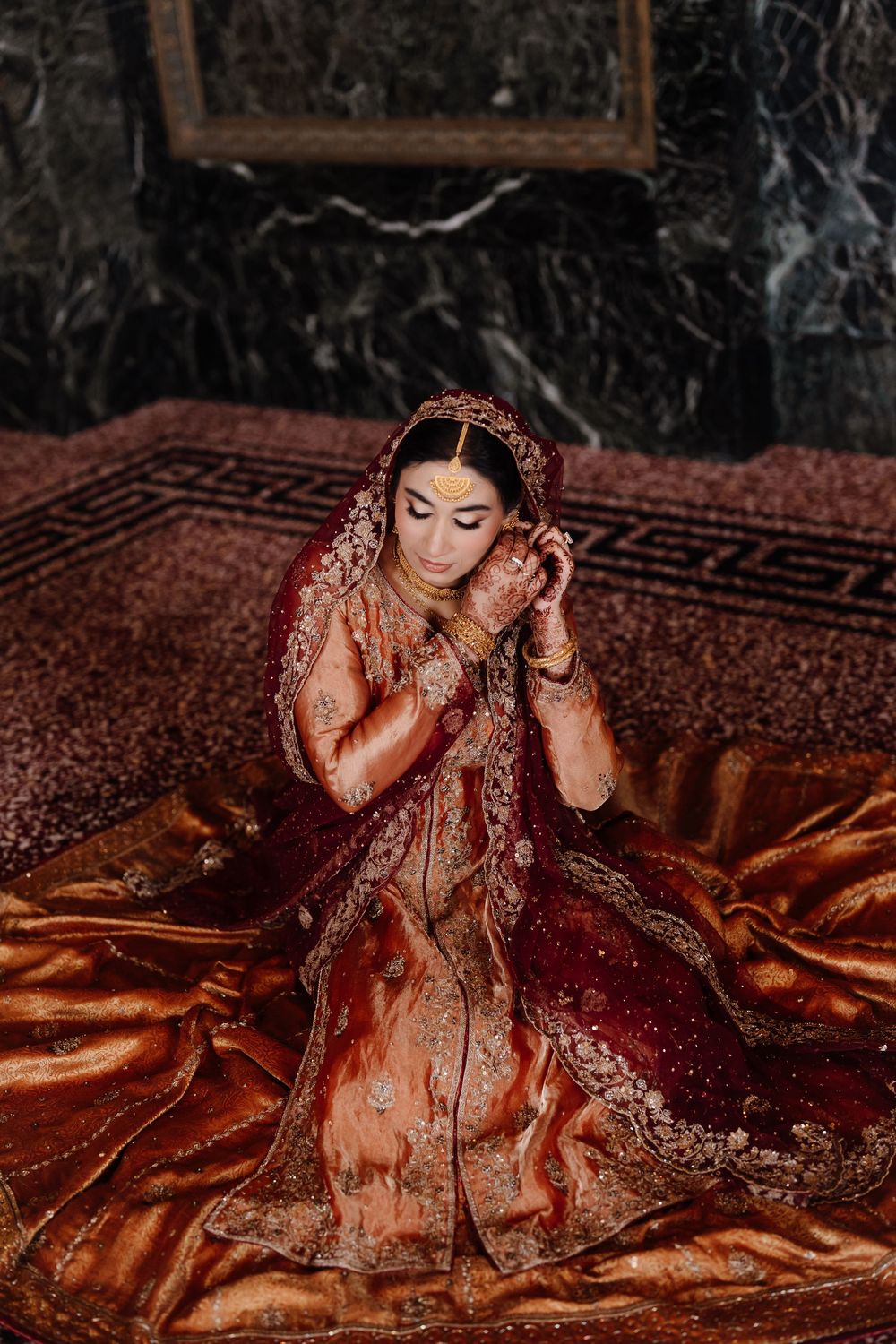 Pakistani bride in elaborate red and gold bridal lehenga adjusting jewelry while seated on ornate carpet.