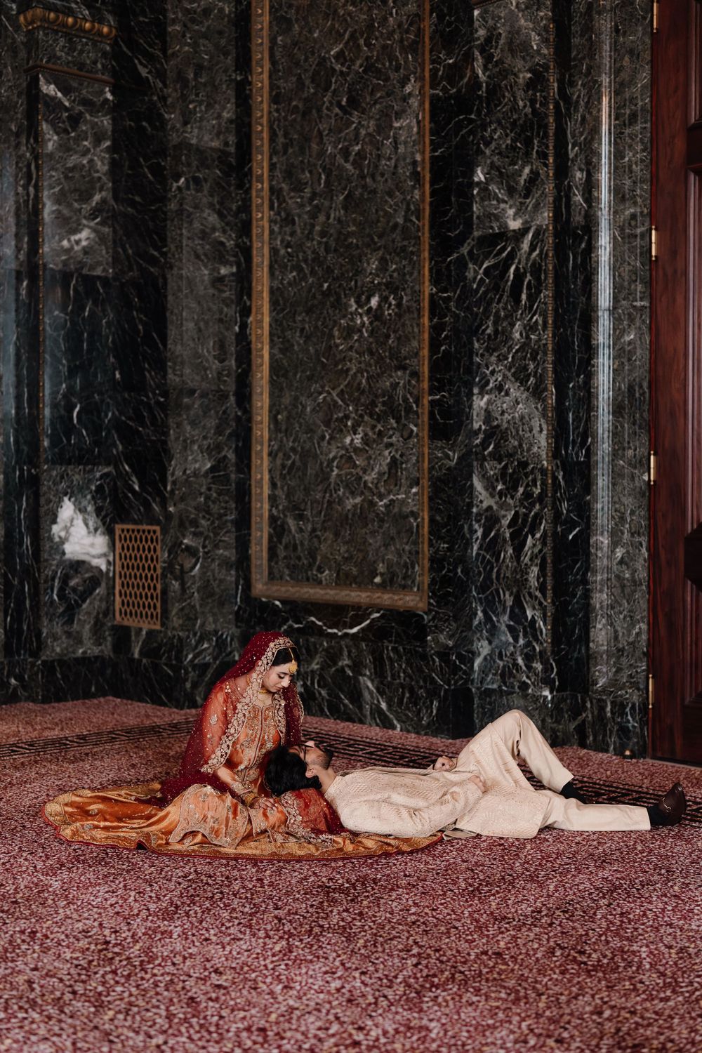 South Asian bride and groom in traditional wedding attire posing on rose petal covered floor against marble walls.
