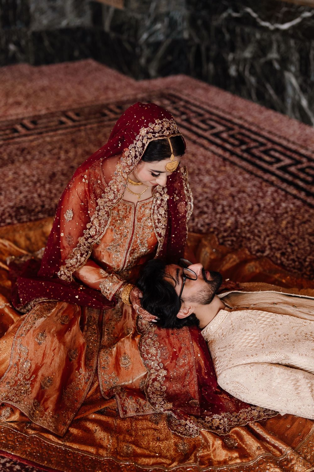 Pakistani bride in red and gold bridal lehenga leaning over groom lying on ornate carpet during wedding photoshoot.