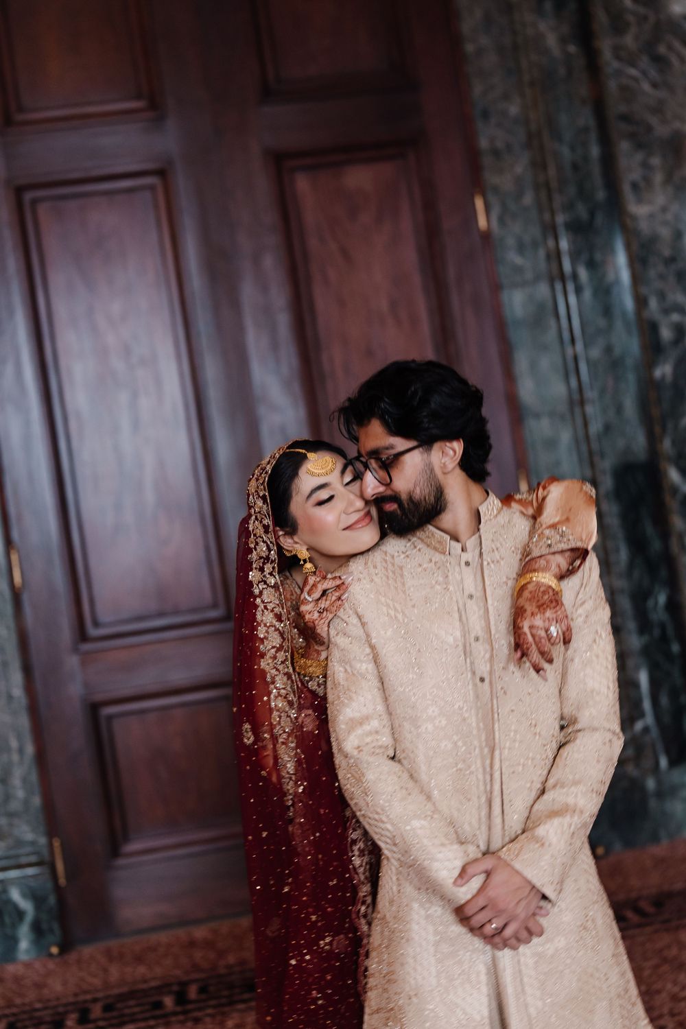 South Asian couple in traditional wedding attire embracing near wooden doors, bride in red dupatta and groom in sherwani.