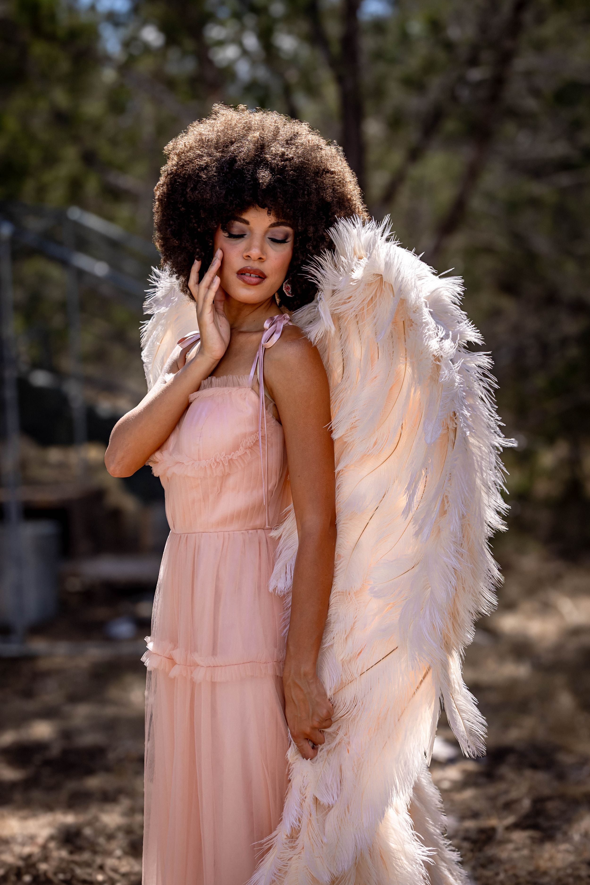 A woman with an afro hairstyle wearing a pink dress and large white feathered angel wings poses outdoors.